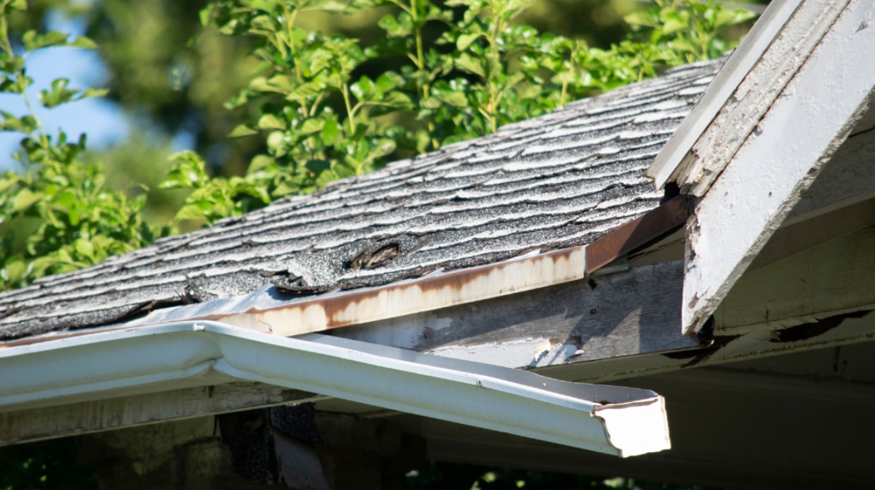 A close-up of a roof's edge showing a rusty gutter and damaged shingles. The roof's condition appears worn and weathered.