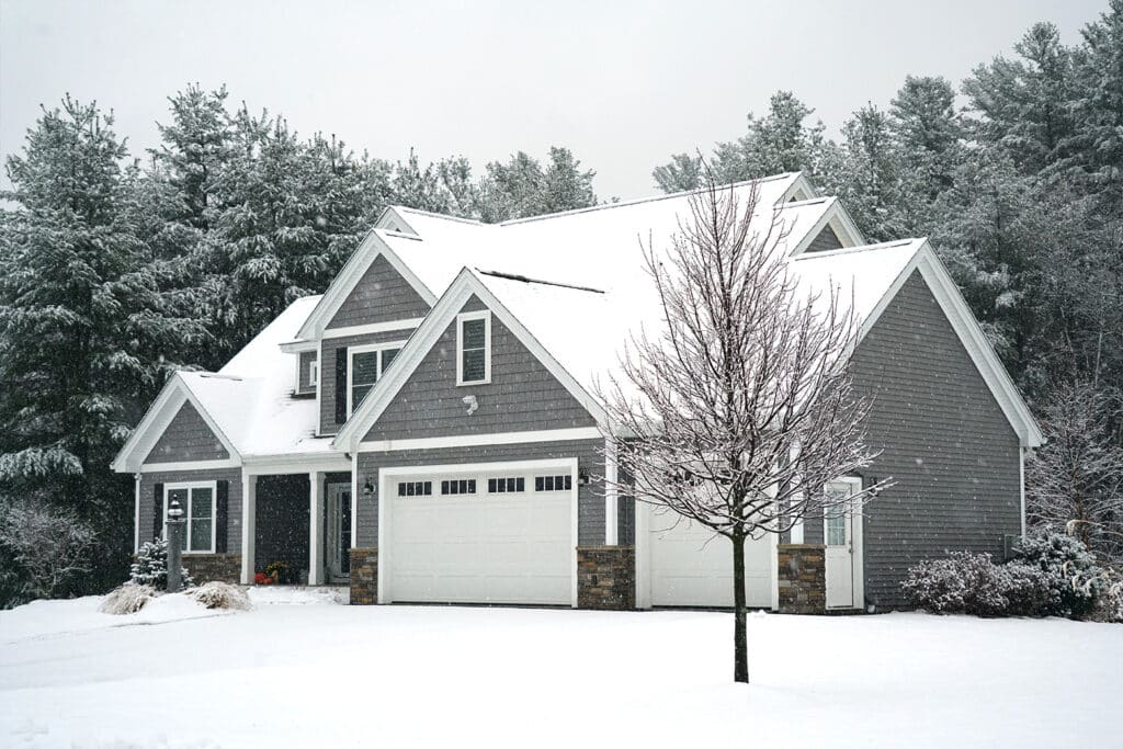 A gray house with white trim and a snow covered roof. The house has a two car garage.