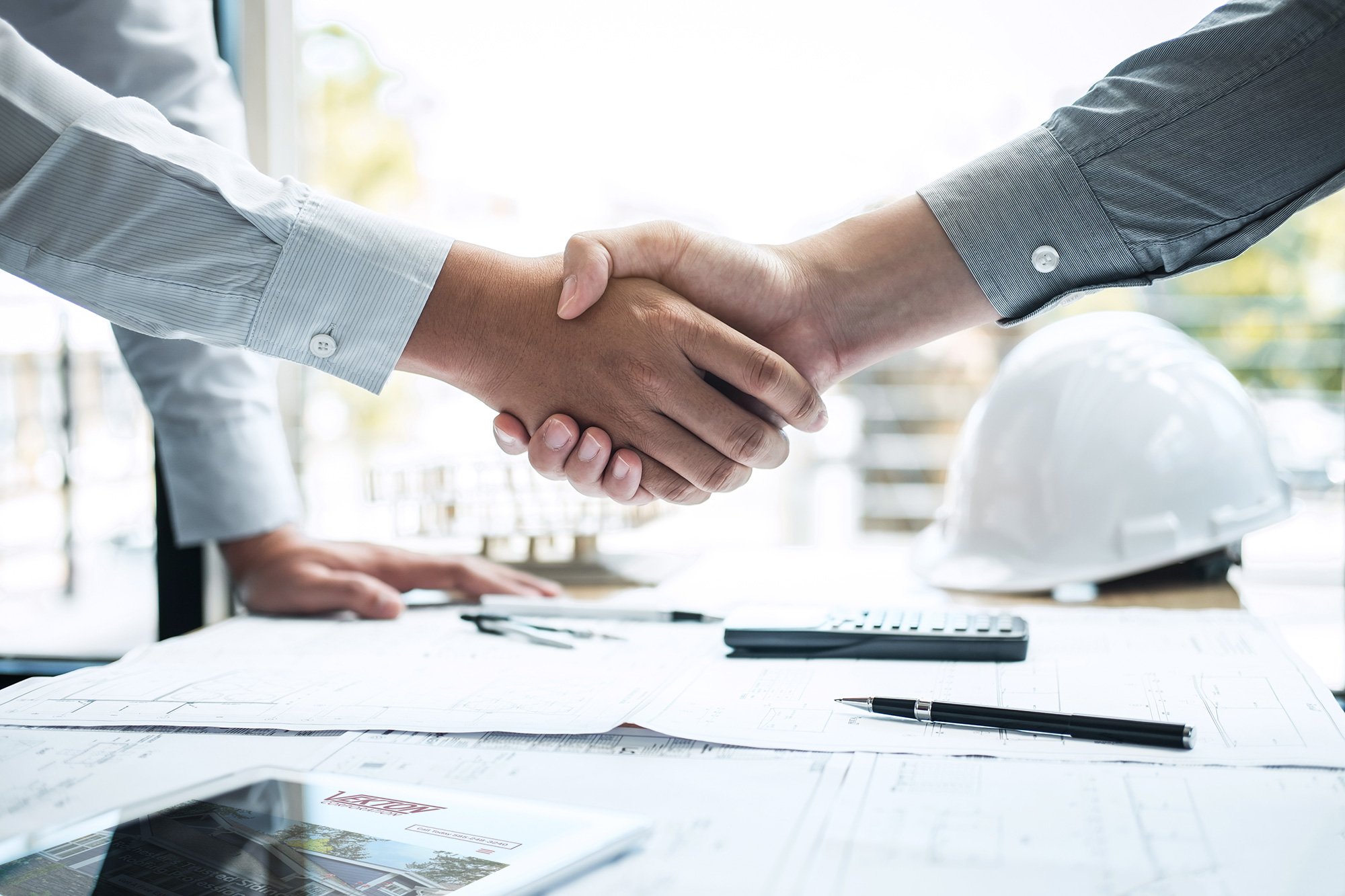 Two businessmen shaking hands. A construction plan and a white hard hat are on the table.