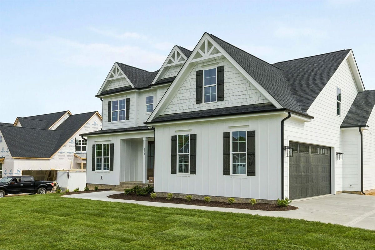 A large white house with a black roof and green shutters. The house has a well-manicured lawn and a driveway leading to a garage.