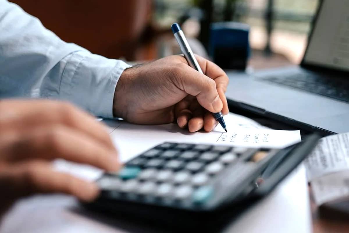 A person is sitting at a desk, using a calculator and pen to calculate numbers on a piece of paper. The person is wearing a light blue shirt and has their hands in focus. A laptop is also visible on the desk. The scene appears to be an office setting.