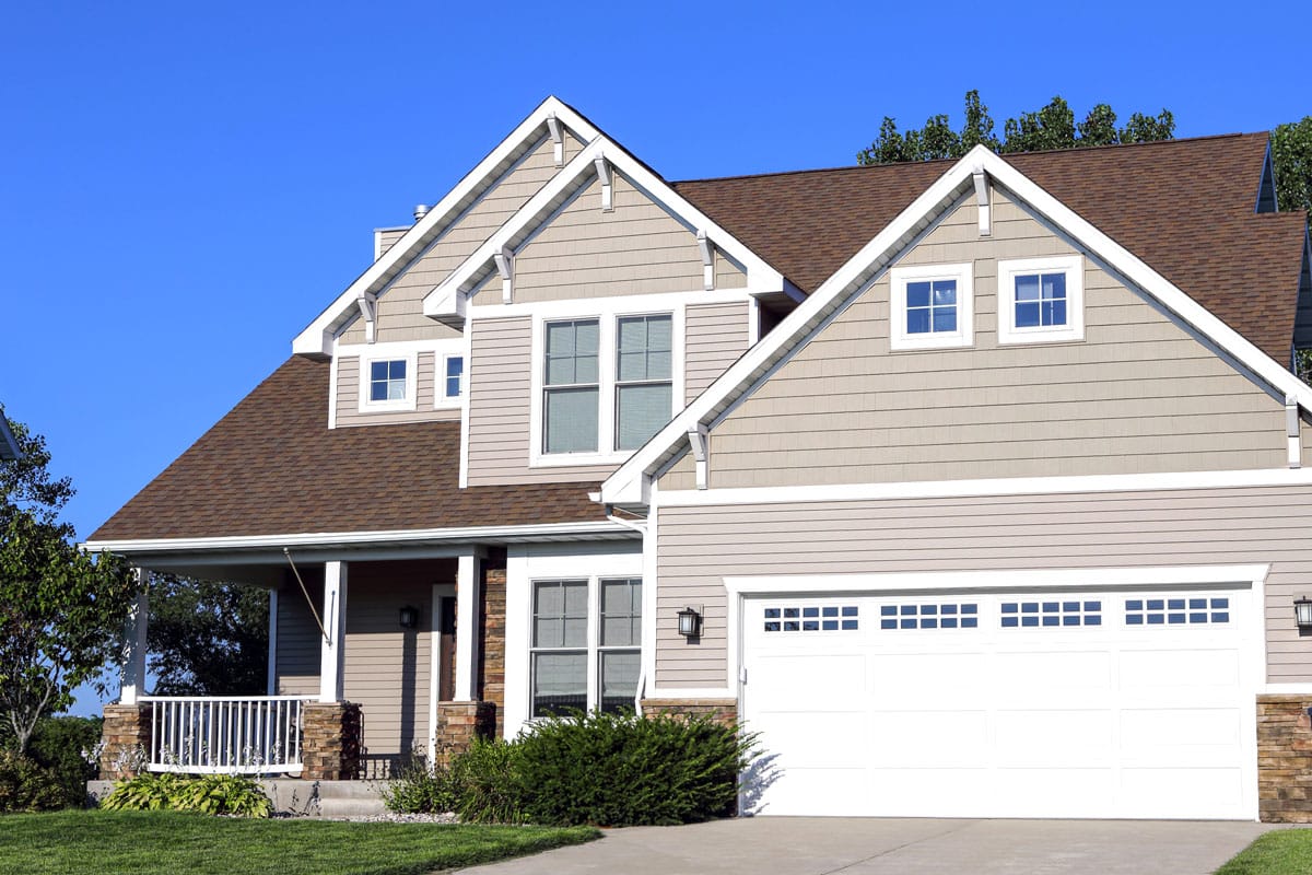 A large two story house with a white garage door and a covered porch. The house has beige siding and a brown roof.