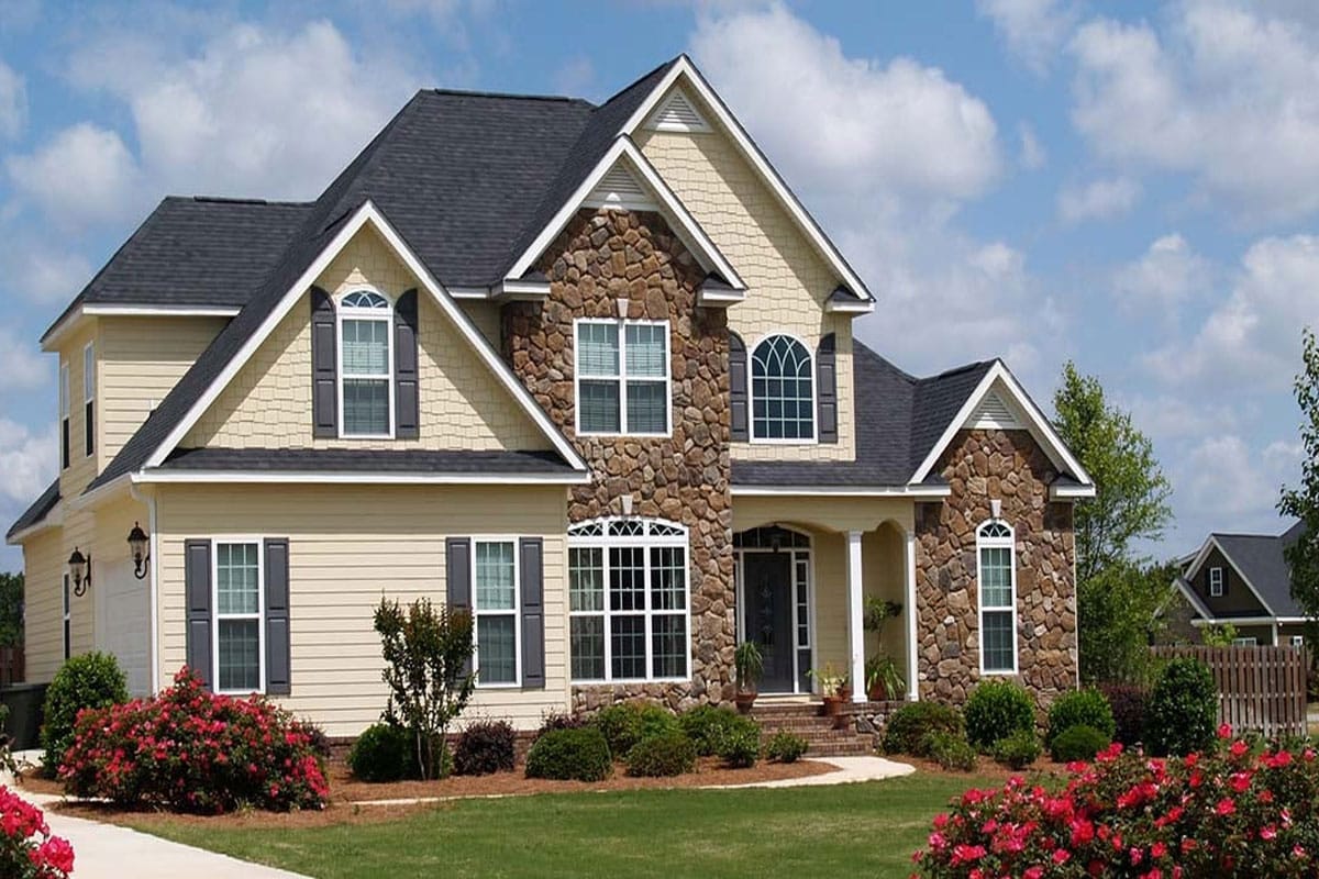 A large two-story house with a stone facade and well-manicured lawn. The house features a black roof and red flowers.