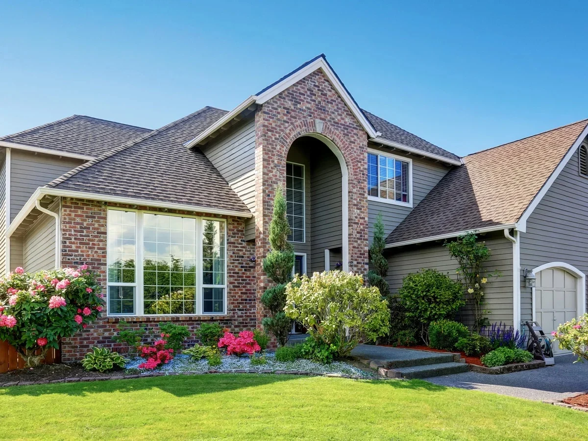 A large house with a brick and vinyl exterior, featuring a large window and a well-manicured lawn. The house has a gray roof and a brick archway.