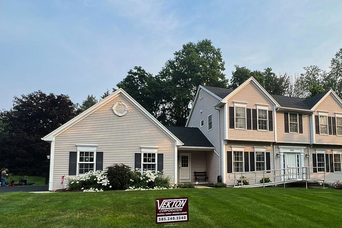 A two-story house with a green lawn and a sign for a roofing company. The house has beige siding and black shutters.