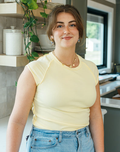 A young woman with a warm smile standing in a kitchen. She is wearing a yellow top and blue jeans.
