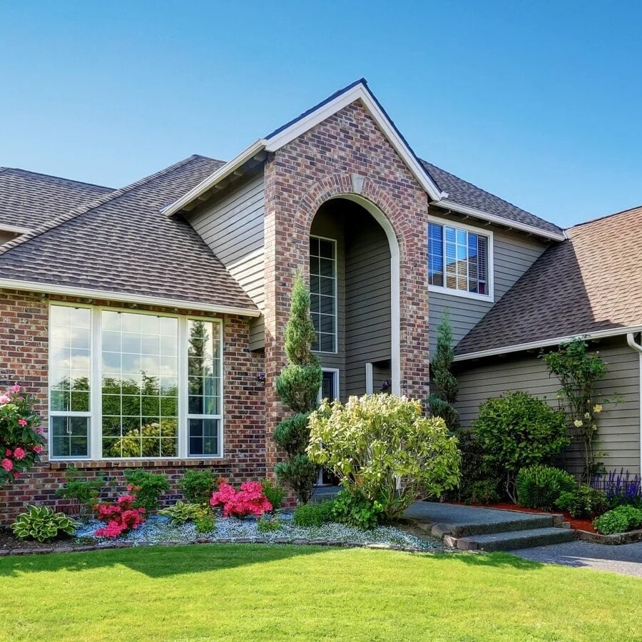 Beautiful Suburban Home with Lush Garden A large house with a brick and vinyl exterior, featuring a large window and a well-manicured lawn. The house has a gray roof and a brick archway.